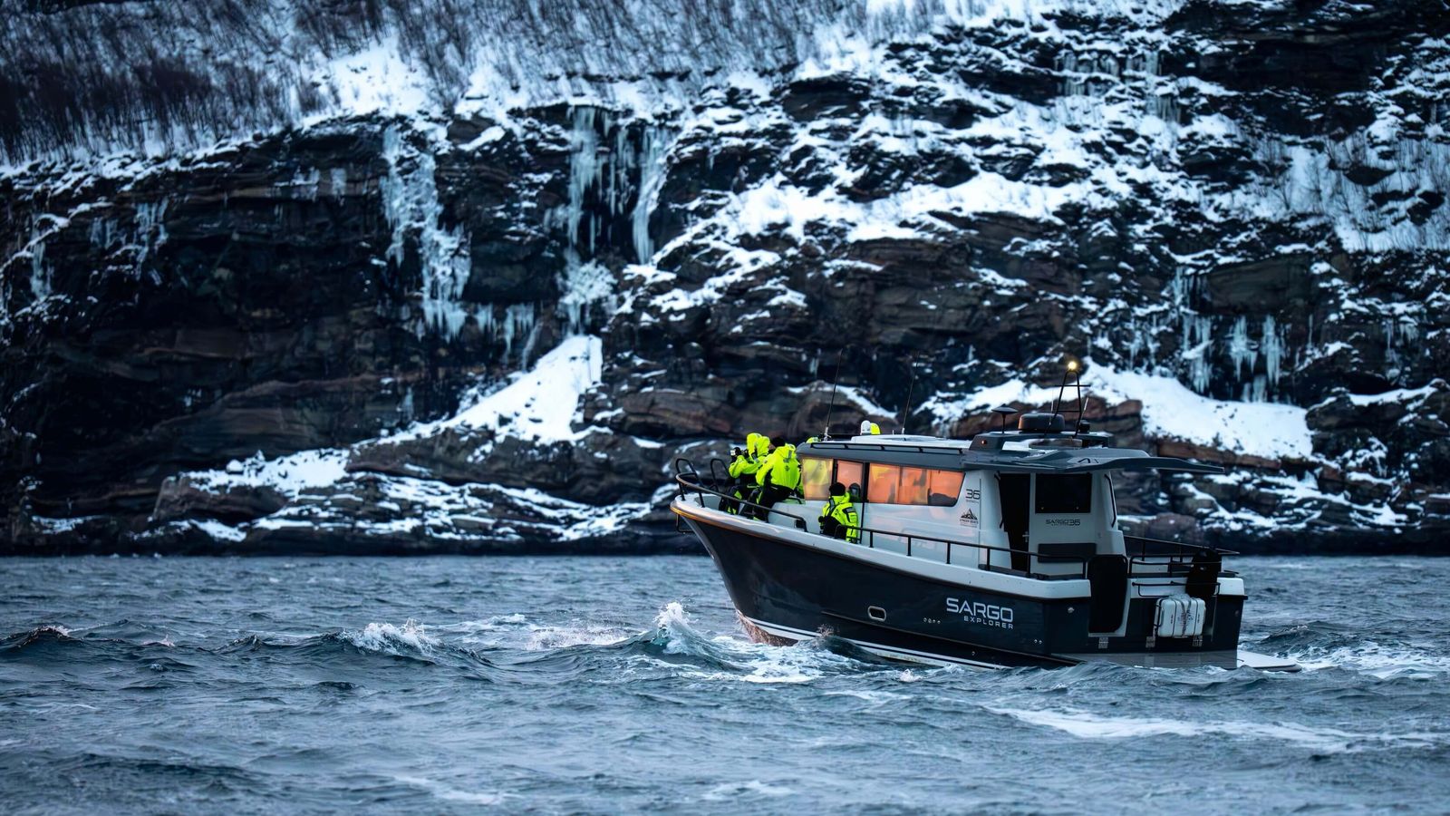 Lyngen Dream med gjester om bord tett under en bratt fjellvegg
